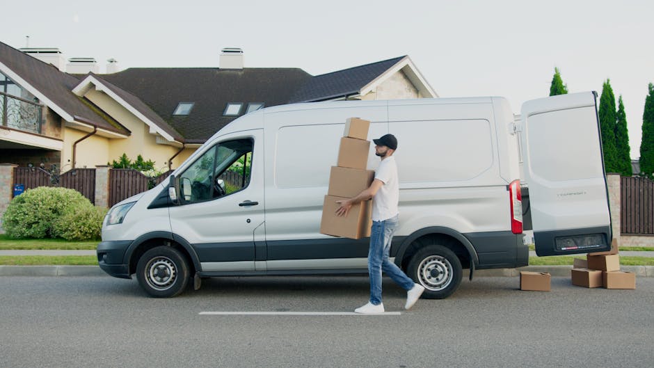 A man wearing a white t-shirt, jeans, and a black cap is carrying three cardboard boxes, stacked one on top of the other, while walking alongside a white cargo van parked on the street in a suburban neighbourhood. The van's rear doors are open, and additional cardboard boxes are placed on the ground next to it. Behind the van, there is a row of modern houses with pitched roofs, large windows, and a variety of plants and bushes along the front yards. The environment appears well-lit with natural daylight, and the scene depicts the process of loading or unloading household items as part of a home relocation or furniture transport service. Man and Van Hounslow offers professional removals, and this image illustrates their loading process in a typical home moving scenario.