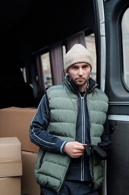 A man stands inside the open sliding door of a moving van, wearing a light grey knit beanie, a green padded sleeveless vest over a navy blue jacket with white stripes on the sleeves, and dark trousers. He is holding a black strap or tie in his gloved hands, ready for securing furniture or boxes during a home relocation. Behind him inside the van, there are cardboard boxes stacked, some wrapped in plastic or packing materials, indicating the packing and moving process. The interior of the van appears to be lined for transport, with visible curtains on the windows. The environment suggests an active loading process, with natural daylight illuminating the scene, relevant to professional removals services like those offered by Man and Van Hounslow.