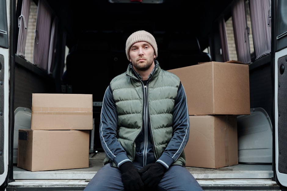 A man wearing a white t-shirt, jeans, and a black cap is carrying three cardboard boxes, stacked one on top of the other, while walking alongside a white cargo van parked on the street in a suburban neighbourhood. The van's rear doors are open, and additional cardboard boxes are placed on the ground next to it. Behind the van, there is a row of modern houses with pitched roofs, large windows, and a variety of plants and bushes along the front yards. The environment appears well-lit with natural daylight, and the scene depicts the process of loading or unloading household items as part of a home relocation or furniture transport service. Man and Van Hounslow offers professional removals, and this image illustrates their loading process in a typical home moving scenario.