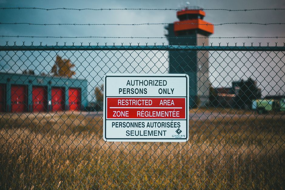 A close-up view of a metal chain-link fence with a sign attached, indicating restricted access for authorized persons only in the Heathrow area. The sign has sections with text in English and French, stating 'AUTHORIZED PERSONS ONLY,' 'RESTRICTED AREA,' and 'PERSONNES AUTORISÉES SEULEMENT.' Behind the fence, there is an industrial setting with large red storage tanks, a control tower or observation building with an orange top, and some parked vehicles. The ground is covered with dry grass, and the weather appears overcast with diffused natural lighting. This scene relates to house removals and moving logistics, as it highlights access restrictions in zones where relocation activities may take place, aligning with the context of managing restricted zones during property and furniture transport in the Heathrow area, as discussed on the [PAGE_TITLE] page of Man and Van Hounslow.