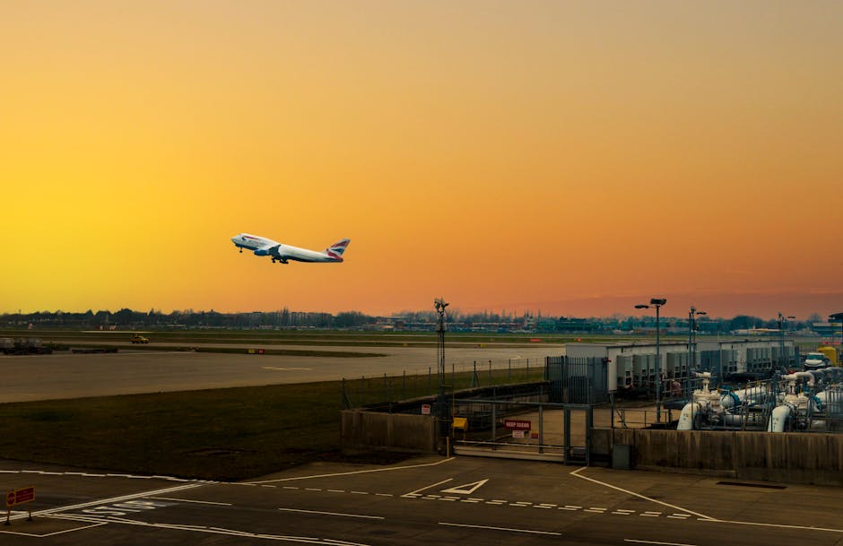 A British Airways commercial airplane is captured mid-air during takeoff above an airport runway at sunset, with a vibrant orange and yellow sky. Below, the airport ground features several industrial-sized fuel tanks, pipelines, and utility structures enclosed within a fenced area. The runway extends into the distance, with small vehicles and airport equipment visible along its length. In the foreground, there are parking bays marked with white lines, and light poles are positioned around the area. The scene demonstrates the typical environment of an international airport, with the airplane preparing for departure amidst various ground facilities. This image relates to transportation and logistics, aligning with house and furniture removals services, such as those provided by Man and Van Hounslow, especially in contexts where precise timing and secure transport are critical for home relocation and furniture transport logistics.