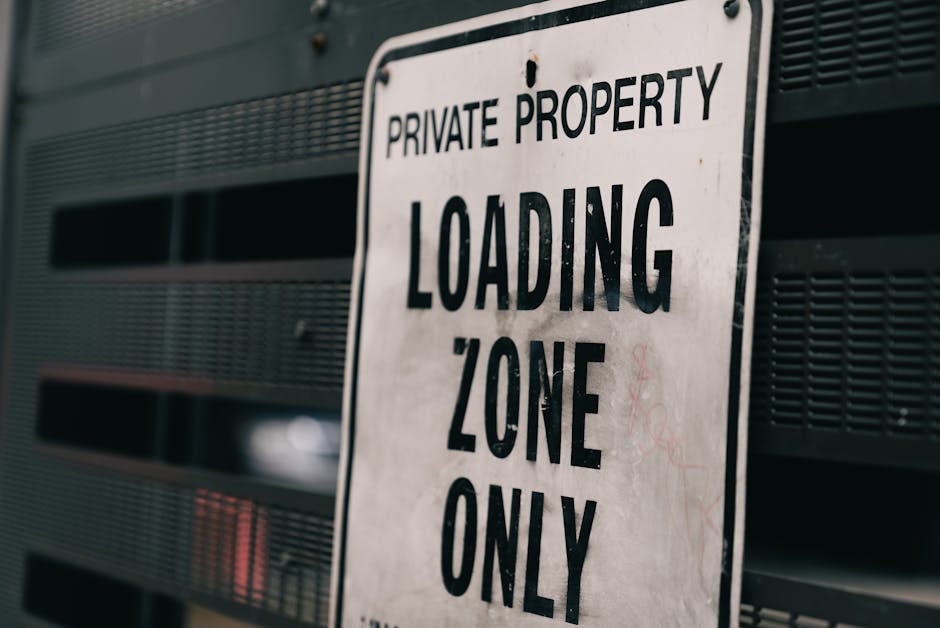 A close-up view of a metal chain-link fence with a sign attached, indicating restricted access for authorized persons only in the Heathrow area. The sign has sections with text in English and French, stating 'AUTHORIZED PERSONS ONLY,' 'RESTRICTED AREA,' and 'PERSONNES AUTORISÉES SEULEMENT.' Behind the fence, there is an industrial setting with large red storage tanks, a control tower or observation building with an orange top, and some parked vehicles. The ground is covered with dry grass, and the weather appears overcast with diffused natural lighting. This scene relates to house removals and moving logistics, as it highlights access restrictions in zones where relocation activities may take place, aligning with the context of managing restricted zones during property and furniture transport in the Heathrow area, as discussed on the [PAGE_TITLE] page of Man and Van Hounslow.
