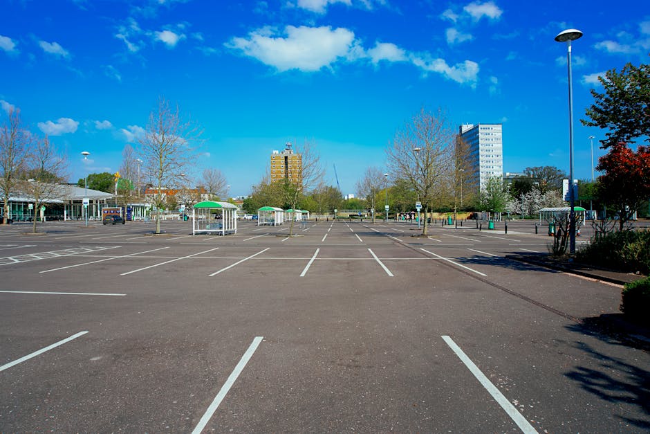 An empty parking lot in Hounslow with multiple marked parking bays, some containing small green and white bus shelters and a few parked cars, set against a backdrop of leafless trees, modern multi-storey buildings, and a clear blue sky with scattered clouds. The lot is well-lit by tall street lamps, and the area appears spacious and organized, suitable for vehicle loading and unloading activities associated with house removals or furniture transport. In this setting, Man and Van Hounslow's removals service can utilize the available parking bays for home relocation logistics, ensuring compliance with local parking permits such as those provided by Hounslow Council for van parking during moving processes.