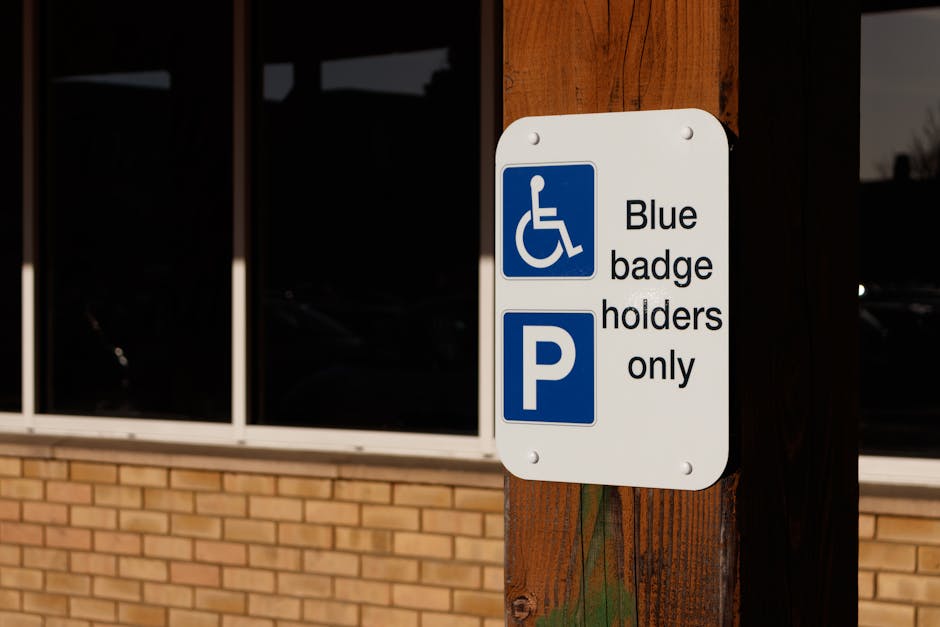 A close-up view of a rectangular white parking sign attached to a wooden post outside a building with large glass windows and a brick lower wall. The sign features a blue wheelchair symbol indicating accessibility, followed by the text 'Blue badge holders only,' and a large blue 'P' symbol below. The sign is positioned near the entrance of the property, which appears to be an office or commercial building. The surrounding environment suggests a parking area designated for vehicles displaying a blue badge, relevant for facilitating home relocation and furniture transport services offered by Man and Van Hounslow. The sign and post are illuminated by natural daylight, emphasizing the clarity of the information displayed, supporting accessible parking for those involved in local removals and moving logistics.
