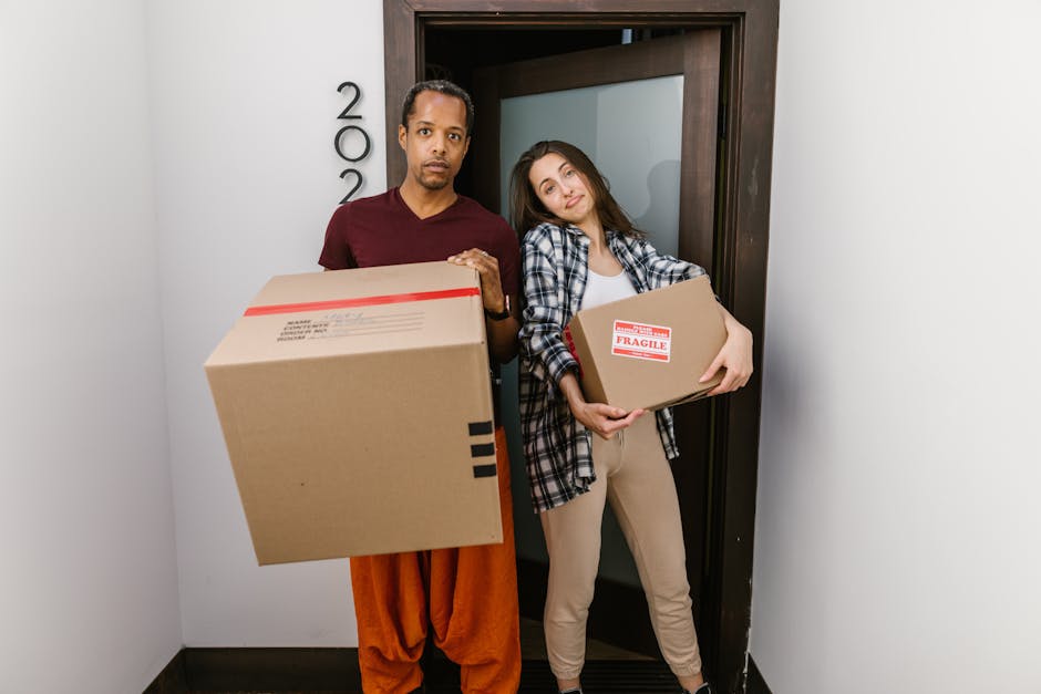 A man and woman stand inside a residential doorway, each holding a cardboard box packed for a house move. The man, dressed in a maroon t-shirt and orange trousers, holds a larger box with black printe