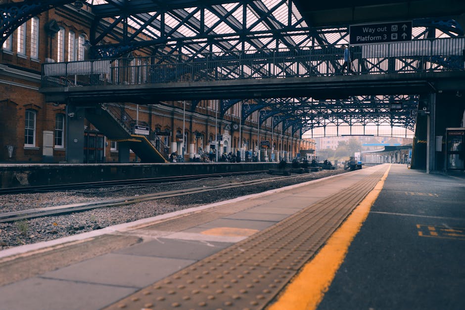 View of a train station platform with a covered metal and glass canopy supported by steel beams and girders. The platform is lined with tactile paving strips in yellow along the edge, and there are a few passengers visible waiting near the station building. The building features brick walls with large windows and staircases on the side leading to an upper level. Railway tracks run parallel to the platform, and a small, dark-colored vehicle can be seen in the distance suggesting ongoing loading or unloading activities. Indoor lighting illuminates the scene, with natural light filtering through the canopy's glass roof. While the image primarily depicts the station environment, it subtly relates to house removals, moving logistics, and the transportation of furniture and boxes through train services, relevant to home relocation considerations. Occasionally, Man and Van Hounslow may utilize such infrastructure for efficient goods transfer during their removals service in the Hounslow area, especially near Hounslow West Station.
