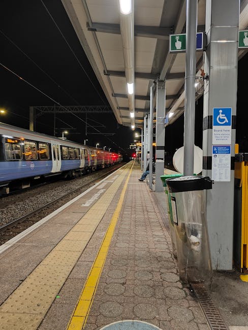 An urban train station platform during daytime with a set of railway tracks running into a tunnel under a brick and metal building. The platform features tactile paving with yellow strips near the edge, and a metal staircase with glass panels and white supports leading up to the station entrance, which is covered with a striped canopy. The station is illuminated by natural light filtering through the tunnel entrance, highlighting the surrounding architecture. The scene provides a neutral view of transit infrastructure relevant to house removals and home relocation logistics, as seen in professional moving services like Man and Van Hounslow.