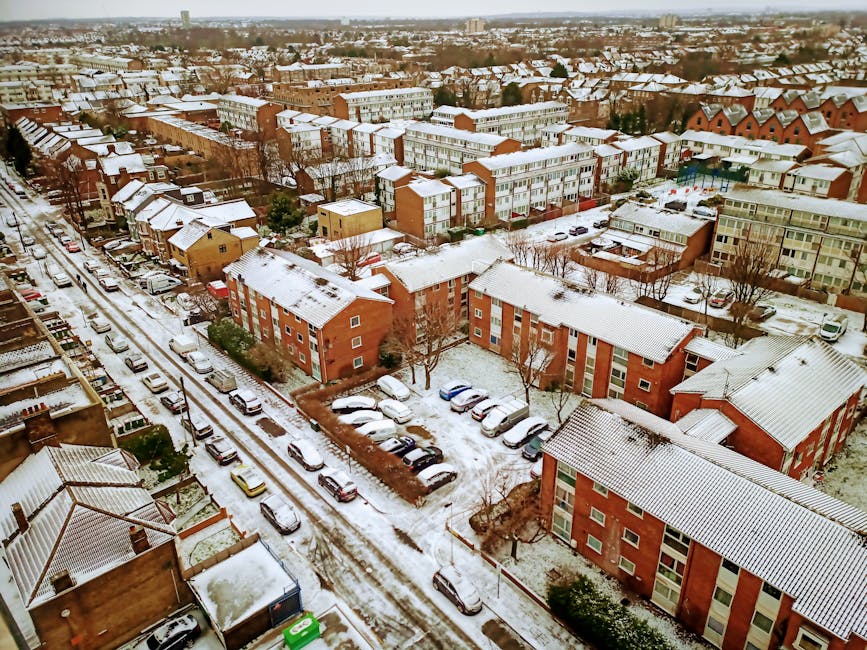 An aerial view of a residential neighbourhood during winter, showing multiple red-brick and cream-colored apartment buildings with snow-covered roofs. The streets are lined with parked cars covered in snow, and a narrow road runs through the area with visible tire tracks. Some cars are parked along the pavement, and leafless trees are scattered throughout the scene. The area appears quiet, with no visible pedestrians or moving vehicles. The sky above is overcast, casting diffuse light over the scene. This image illustrates an urban setting ideal for home relocation or furniture transport services, aligning with the offerings of Man and Van Hounslow for same-day removals and moving logistics within the area.