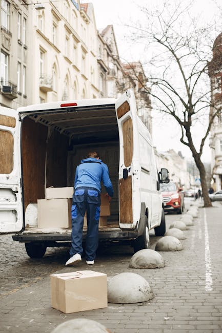 A man dressed in a blue jacket and dark trousers is seen loading cardboard boxes into the rear of a white moving van parked on a city street. The back doors of the van are open, revealing the interior lined with wooden panels, and he is placing a box inside on the floor. Additional packed boxes are positioned on the pavement nearby, some stacked against the curb. The street is lined with multi-story buildings featuring ornate facades, and a leafless tree stands along the sidewalk. Several parked cars are visible along the curb, and concrete bollards separate the pedestrian walkway from the roadway. The scene depicts a typical home relocation or furniture transport process carried out by a removals company, fitting the context of packing and moving services, with natural daylight and an overcast sky providing neutral lighting.