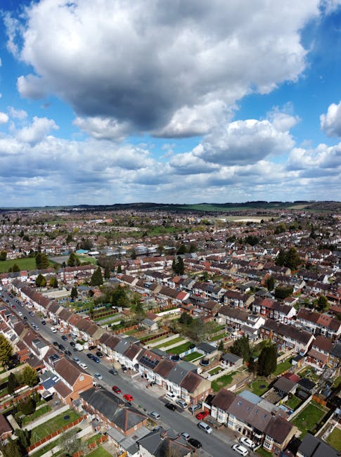 An aerial view of a residential neighbourhood during winter, showing multiple red-brick and cream-colored apartment buildings with snow-covered roofs. The streets are lined with parked cars covered in snow, and a narrow road runs through the area with visible tire tracks. Some cars are parked along the pavement, and leafless trees are scattered throughout the scene. The area appears quiet, with no visible pedestrians or moving vehicles. The sky above is overcast, casting diffuse light over the scene. This image illustrates an urban setting ideal for home relocation or furniture transport services, aligning with the offerings of Man and Van Hounslow for same-day removals and moving logistics within the area.