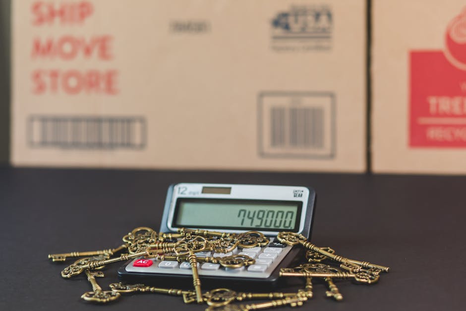 A calculator displaying the number 749800 is positioned on a black surface, surrounded by numerous antique brass keys of various sizes and shapes, some with ornate bow designs, spread in front of a background of cardboard boxes with red and blue labels. The boxes are stacked, with some visible text indicating shipping, moving, and store instructions. The scene appears to be set in a storage or packing area, with the keys and calculator suggesting a focus on the valuation or inventory process related to house removals and home relocation. The lighting is even and neutral, emphasizing the objects' details without shadows or glare. This visual context reflects the careful handling, packing, and logistics involved in furniture transport and moving services, as offered by companies like Man and Van Hounslow, supporting topics around removal costs, packing and moving, including the intricacies of property and furniture transportation.