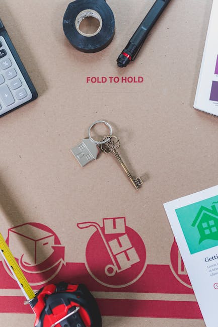 An overhead view of a workspace related to home relocation, featuring a cardboard moving box with red printed instructions for folding and holding, partially visible at the edges. On the box, there are various packing materials, including a roll of black adhesive tape, a black marker pen, and a white tablet displaying green and purple graphics. Near the center, a silver house-shaped keyring with a small metal key and a tiny grey plastic house charm is placed on the box. To the left, part of a grey calculator is visible, and below, a yellow measuring tape extends across the surface. The background surface appears to be a plain beige table or countertop. This scene exemplifies the packing process in house removals, with tools and packaging materials ready for organized furniture transport, supported by the professional removals services of Man and Van Hounslow.